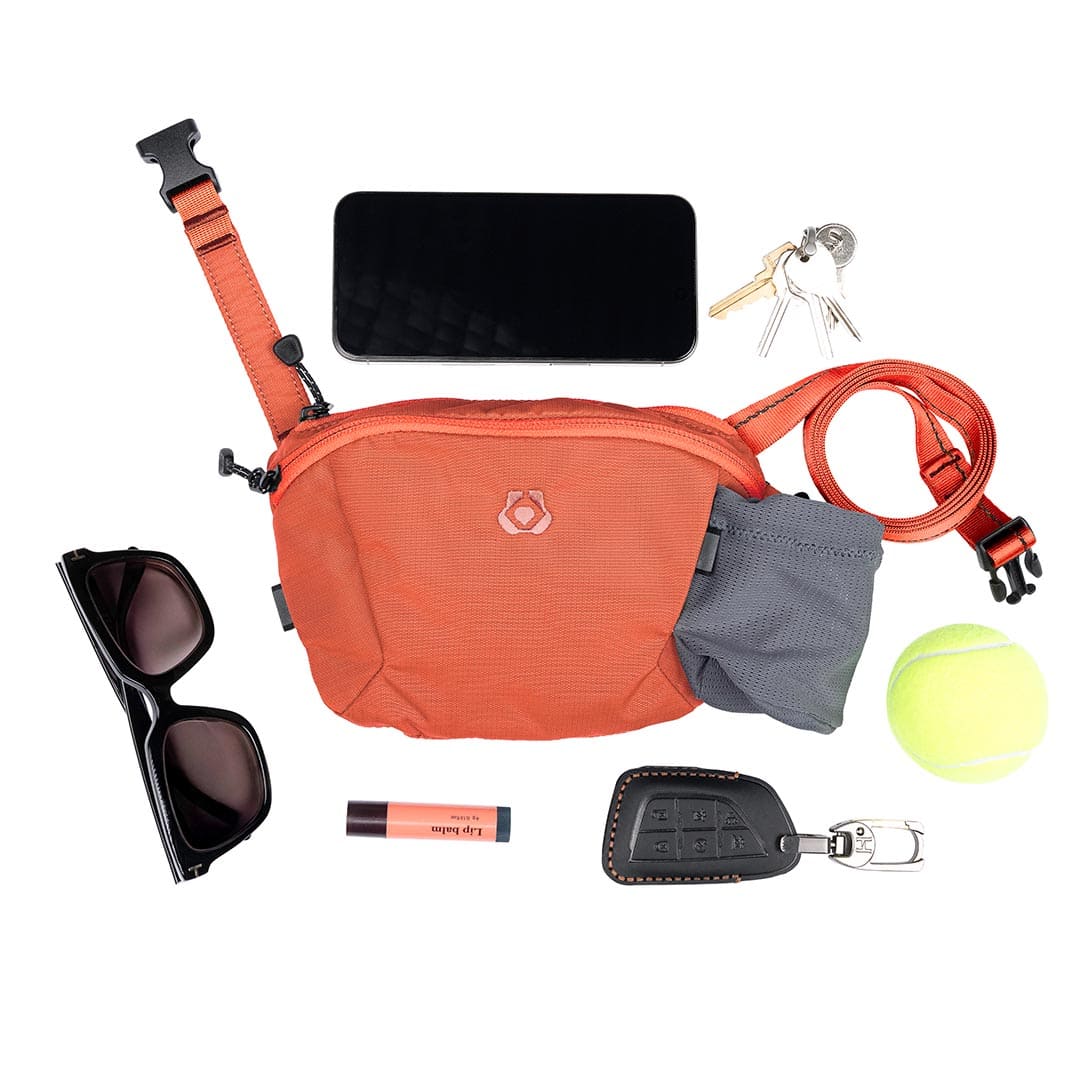 Orange waist bag with various items including sunglasses, phone, keys, dog leash, and tennis ball on a white background